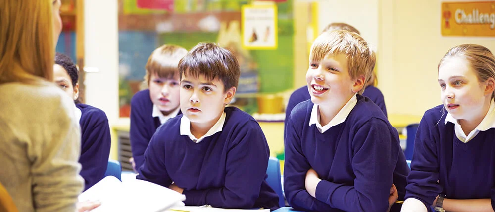 A group of children in school uniforms sit attentively in a classroom, listening to a teacher who is partially visible.