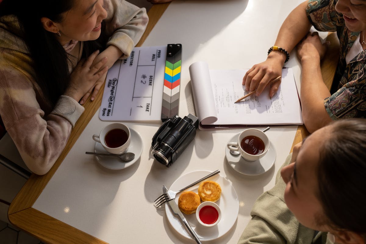 Three people sit at a table with a clipboard, notebook, video camera, two cups of tea, and pastries, engaged in discussion and note-taking as they plan pre-production for a scripted video.