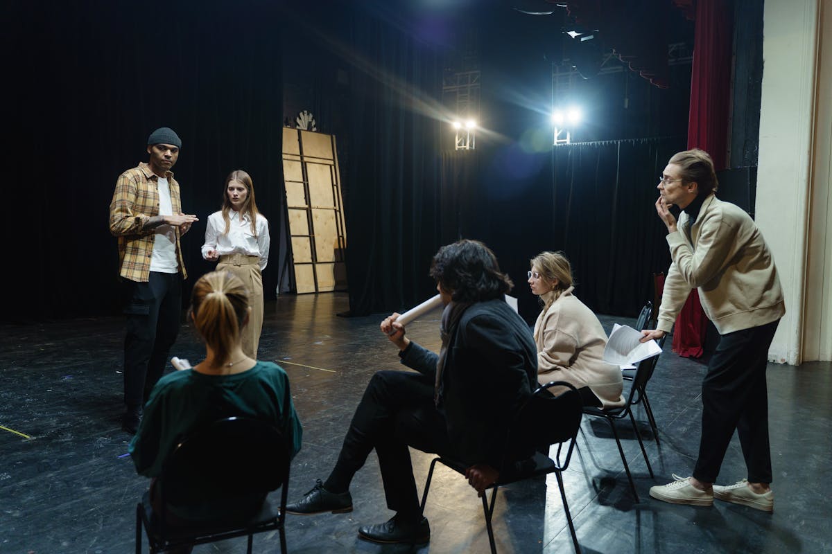 A group of people rehearses on a dimly lit theater stage; two stand facing four others who are seated and watching attentively.