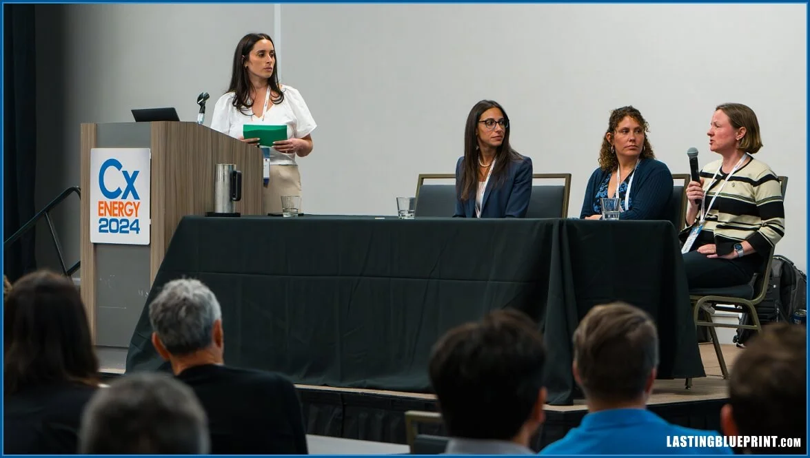 Four women seated at a panel discussion at the cx energy 2024 conference. One woman stands at a podium labeled "cx energy 2024," while another speaks into a microphone. Audience members are visible in the foreground.