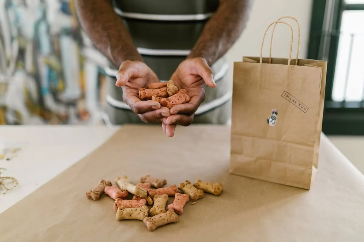 A person holding various dog biscuits in their hands over a table with more biscuits and a brown paper bag.