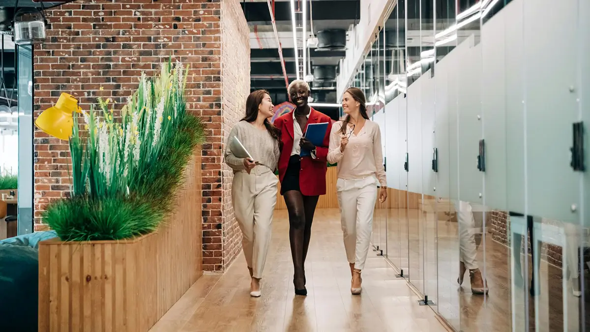 Three colleagues walk down a modern office hallway with a brick wall and glass partitions, engaging in conversation. The person in the red blazer holds a blue folder.
