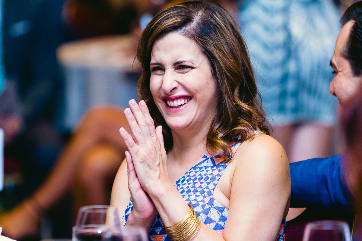 A woman in a blue and white patterned dress is clapping and smiling while seated at an event.