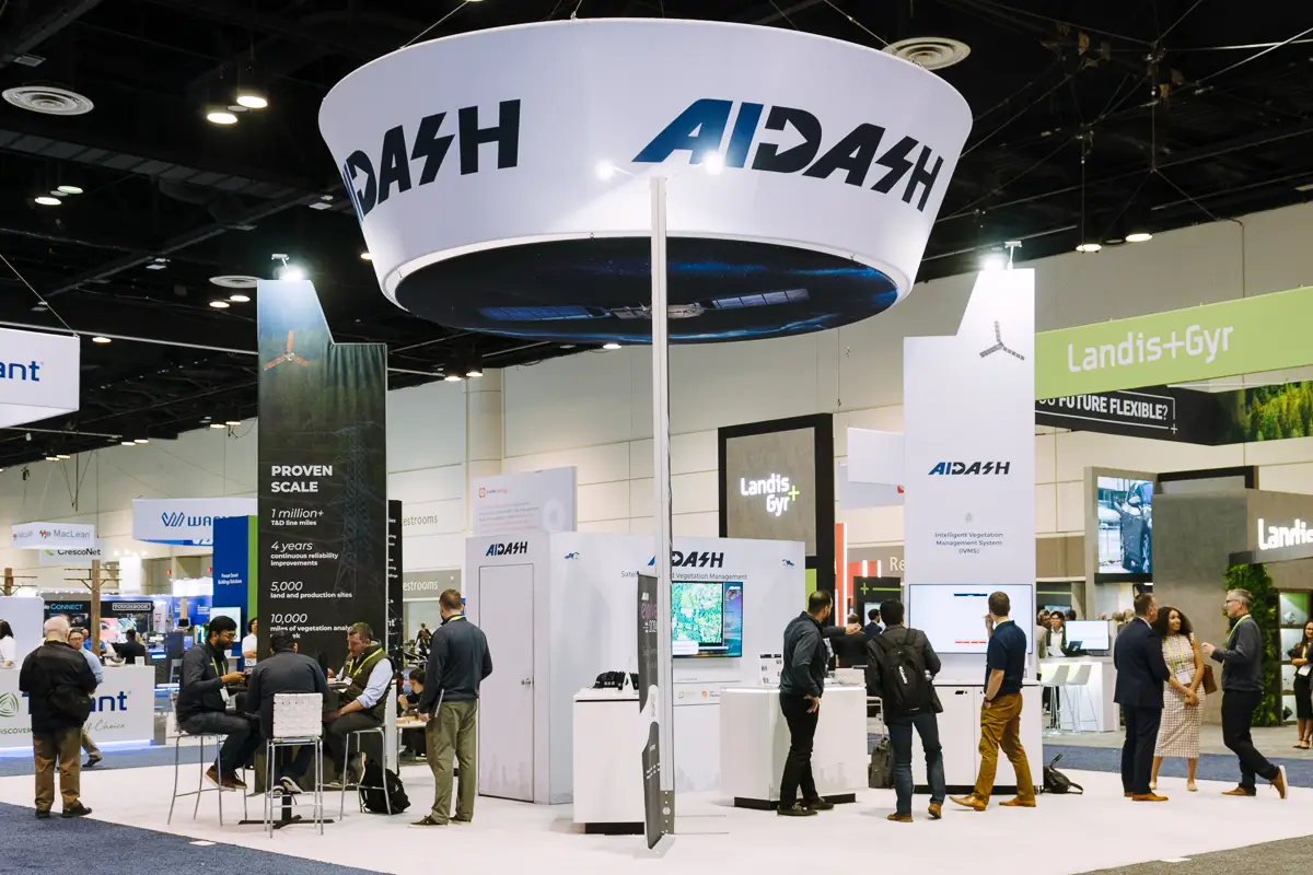 People walk around visiting booths at a tech conference at the orange county convention center. A prominent circular sign hangs above the aidash booth.