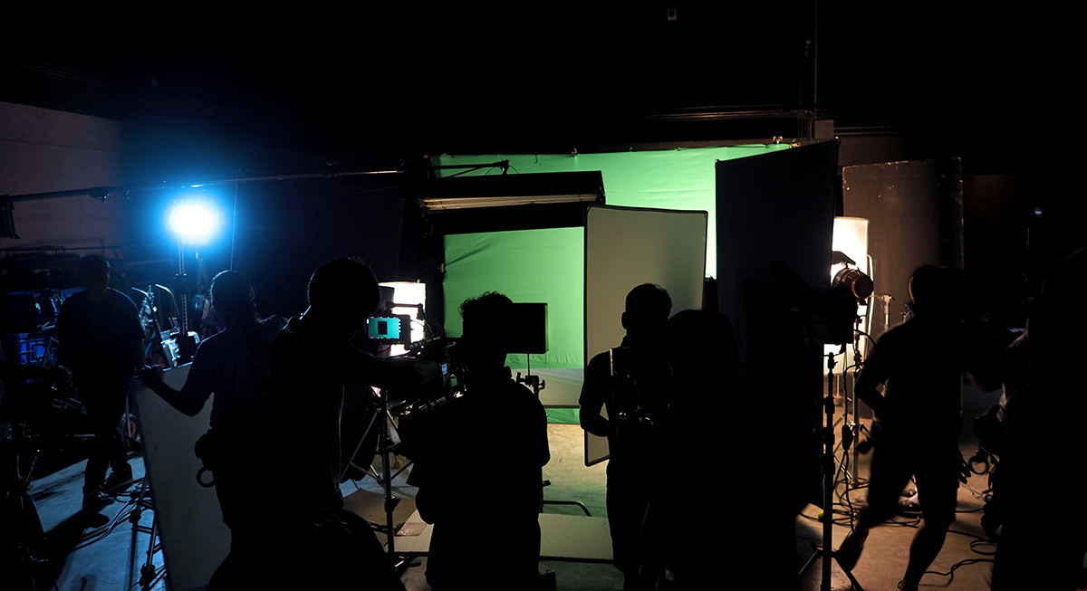 A dimly lit film set with crew members silhouetted against bright studio lights and a green screen background captures the intricate video production stages.