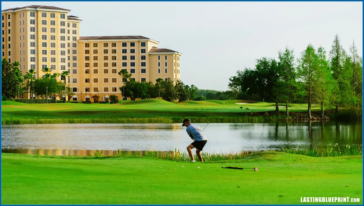 A golfer in mid-swing stands near a pond with a lush green golf course and a tall hotel building in the background.