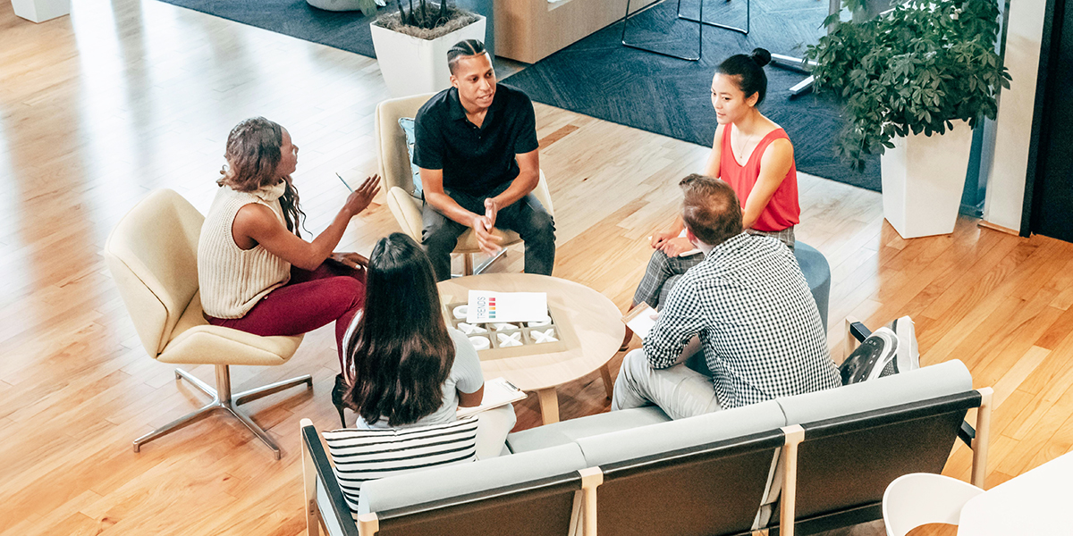 A group of people seated in a circle engages in discussion in a modern office with plants and large windows.