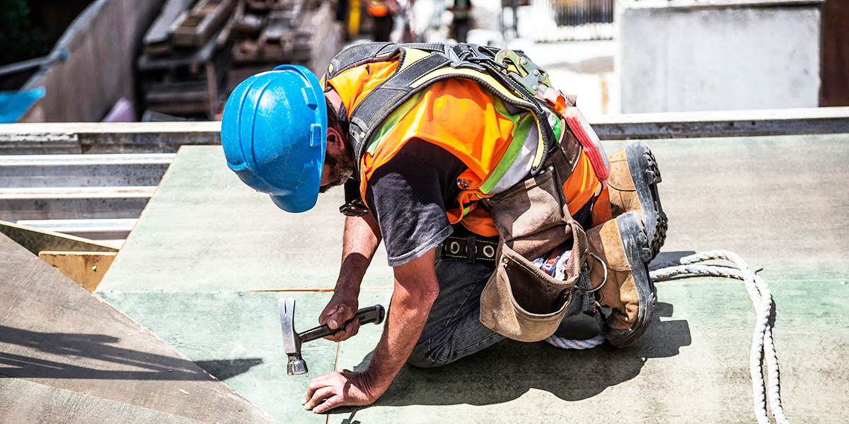 A construction worker in safety gear kneels on a structure, hammering a nail into the surface of the bustling orlando construction project.