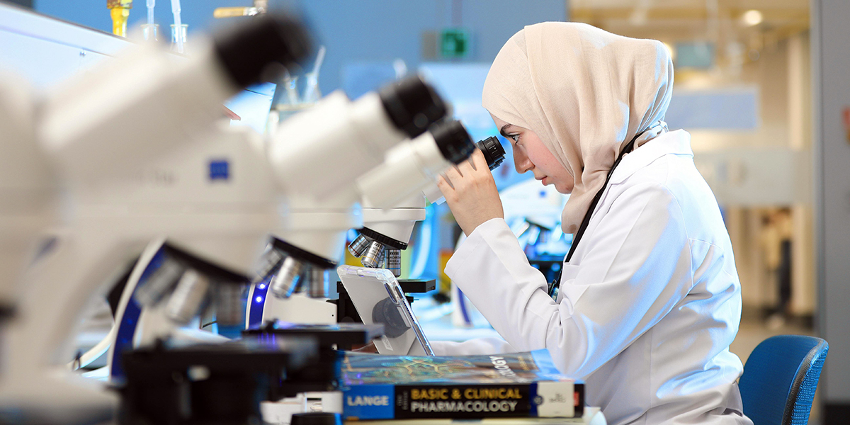 A woman in a lab coat and hijab looks into a microscope at a laboratory bench with textbooks and other microscopes nearby.