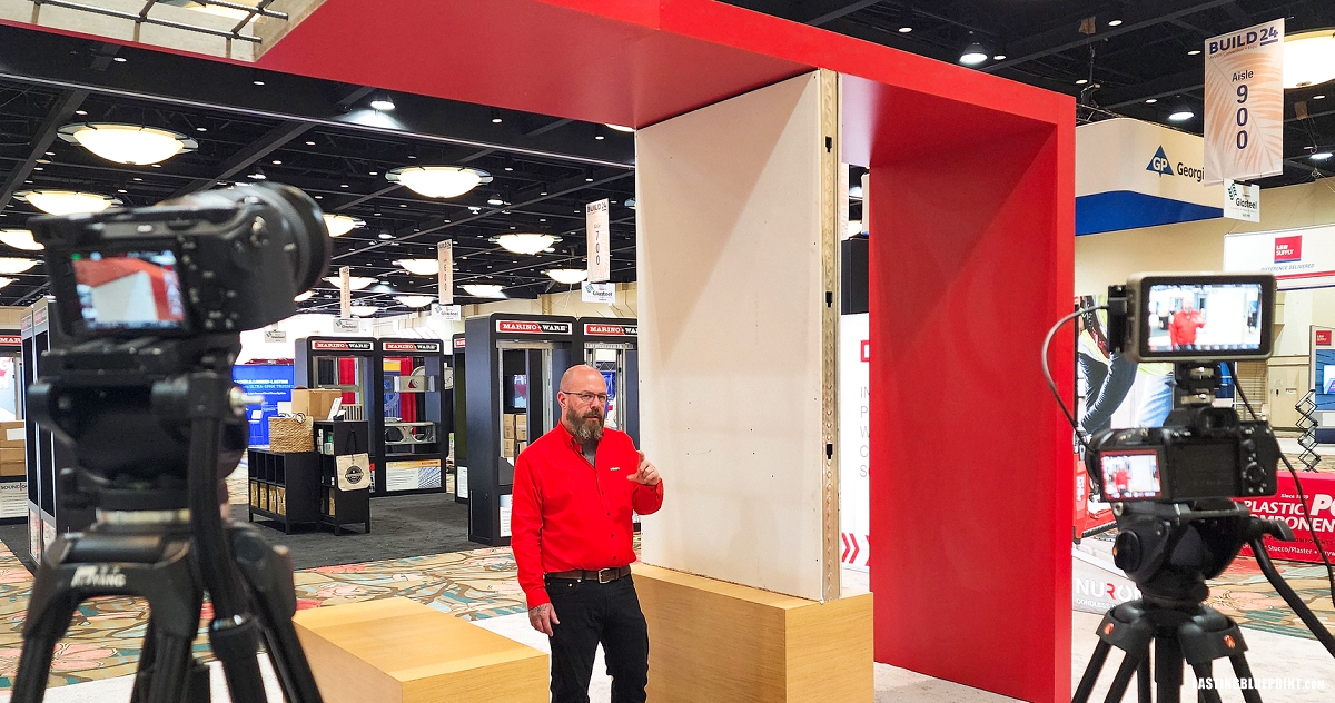 A man in a red shirt stands between two cameras, presenting at a trade show booth with displays and signage in the background at the walt disney world swan & dolphin