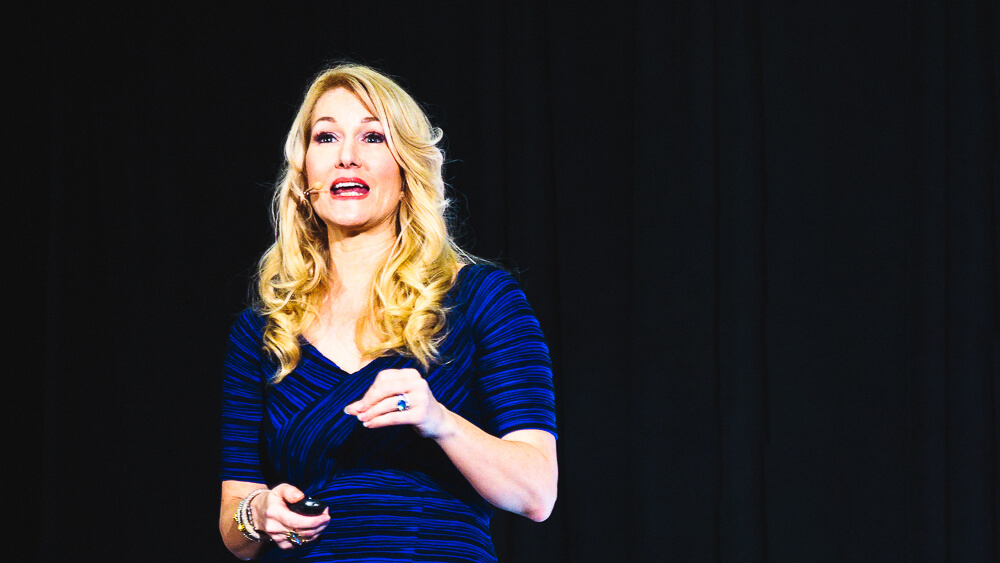 At the orlando conference, a videographer captures a person with long blonde hair in a blue dress speaking on stage against a dark background.